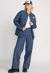 Blaue Baumwolljacke mit einem braunen Kragen, kombiniert mit passenden blauen Hosen und einer blauen Mütze mit einem Logo. Schwarze Plateaustiefel.