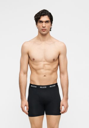 Homme torse nu aux cheveux foncés portant un boxer noir Hollister, debout devant un fond clair uni, faisant face à l'avant.