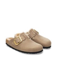 Sabots en suède beige avec deux sangles réglables et des boucles dorées. Doublés de fourrure beige pour le confort. Semelles en caoutchouc noir pour une meilleure traction.