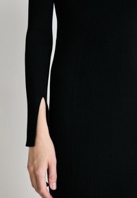Black ribbed dress featuring long sleeves with a slit at the wrist, showcasing a fitted design and textile texture.