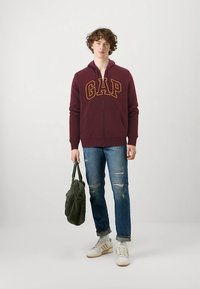 Vinrød hættejakke med lynlås og "GAP"-logo, denimjeans med slidte detaljer, hvide sneakers og grøn taske. Skarpt, afslappet outfit.