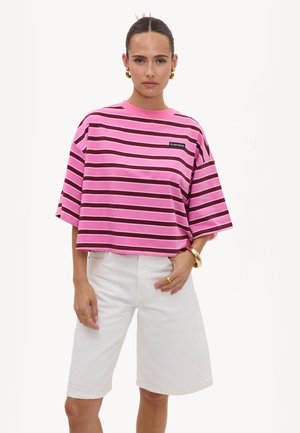 Femme portant un t-shirt oversize rayé rose et violet avec un short blanc longueur genou, des boucles d'oreilles créoles dorées et des bijoux en or sur fond blanc.