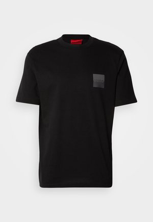 T-shirt en coton noir à col rond, avec un patch texturé noir portant un logo sur la poitrine gauche. Manches courtes et coupe décontractée.