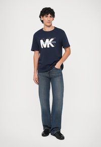Joven vistiendo una camiseta de Michael Kors azul marino, jeans azules y zapatillas negras, de pie con una mano en el bolsillo frente a un fondo liso.
