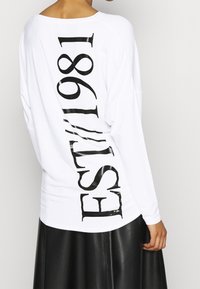 Person trägt ein weißes Langarmshirt mit großen vertikalen "EST. 1981"-Schriftzug in Schwarz auf dem Rücken und einen schwarzen Lederrock.