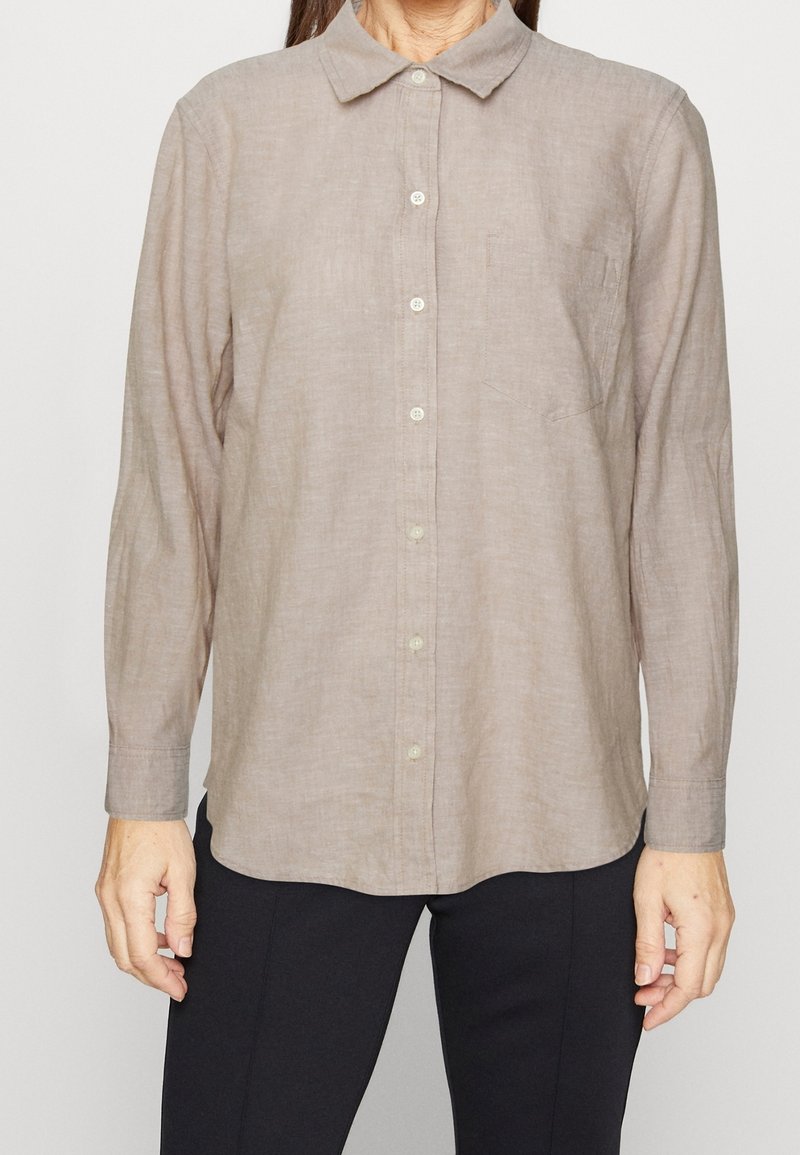 Femme portant une chemise marron clair à manches longues boutonnée avec col et poche poitrine, associée à un pantalon noir, debout devant un fond uni.