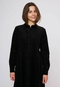 Rochie cambrată din cotele negre, cu mâneci lungi, închidere cu nasturi în față și fustă evazată. Materialul are un aspect moale și texturat.