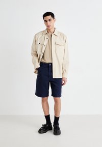 Beige jacket over a beige t-shirt, navy shorts, black socks, and black shoes. The jacket features two front pockets and a relaxed fit.