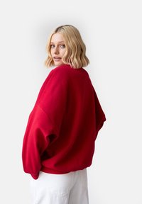 Czerwona oversize'owa bluza z miękką teksturą i ściągaczami, w zestawieniu z białymi spodniami. Projekt ma okrągły dekolt i luźny krój.