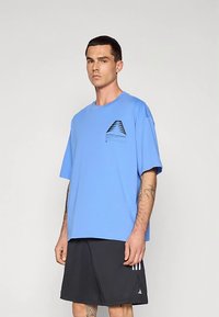 T-shirt à manches courtes bleu clair avec un motif géométrique noir sur le côté gauche de la poitrine, associé à un short gris foncé avec trois bandes.