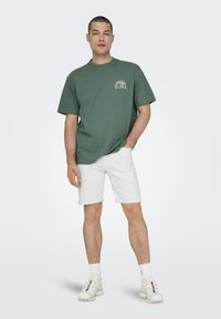 Camiseta de algodón verde con mangas cortas y un estampado gráfico en el pecho, combinada con pantalones cortos de mezclilla blancos enrollados y zapatillas blancas.