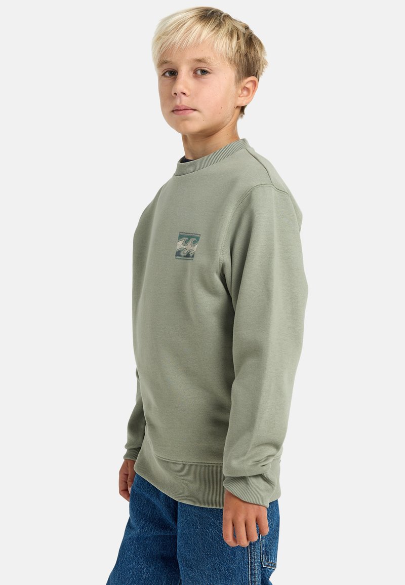 Felpa verde con colletto rotondo, polsini e orlo a costine. Presenta una piccola grafica a forma di onda in blu e crema sulla parte anteriore. Jeans in denim.