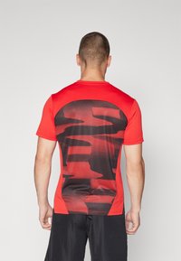 Camiseta atlética roja con un patrón de camuflaje a rayas horizontales negras en la parte posterior. Diseño de mangas cortas y cuello redondo, hecha de una tela suave.