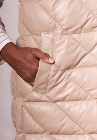 Hand with dark skin tone resting inside the side pocket of a beige quilted leather jacket.