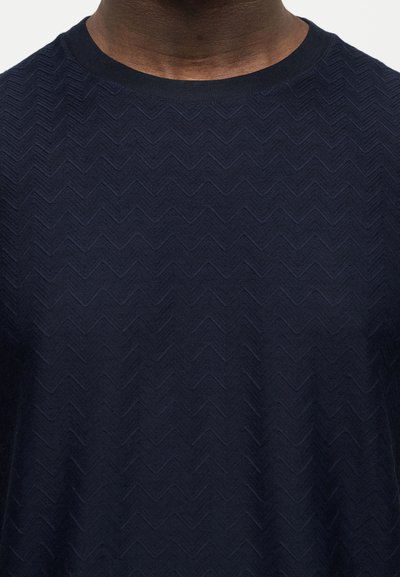 Άτομο με σκουρόχρωμο δέρμα που φοράει πουλόβερ πλεκτό σε σκούρο μπλε, με υφή και μοτίβο ψαροκόκκαλο και στρογγυλή λαιμόκοψη.