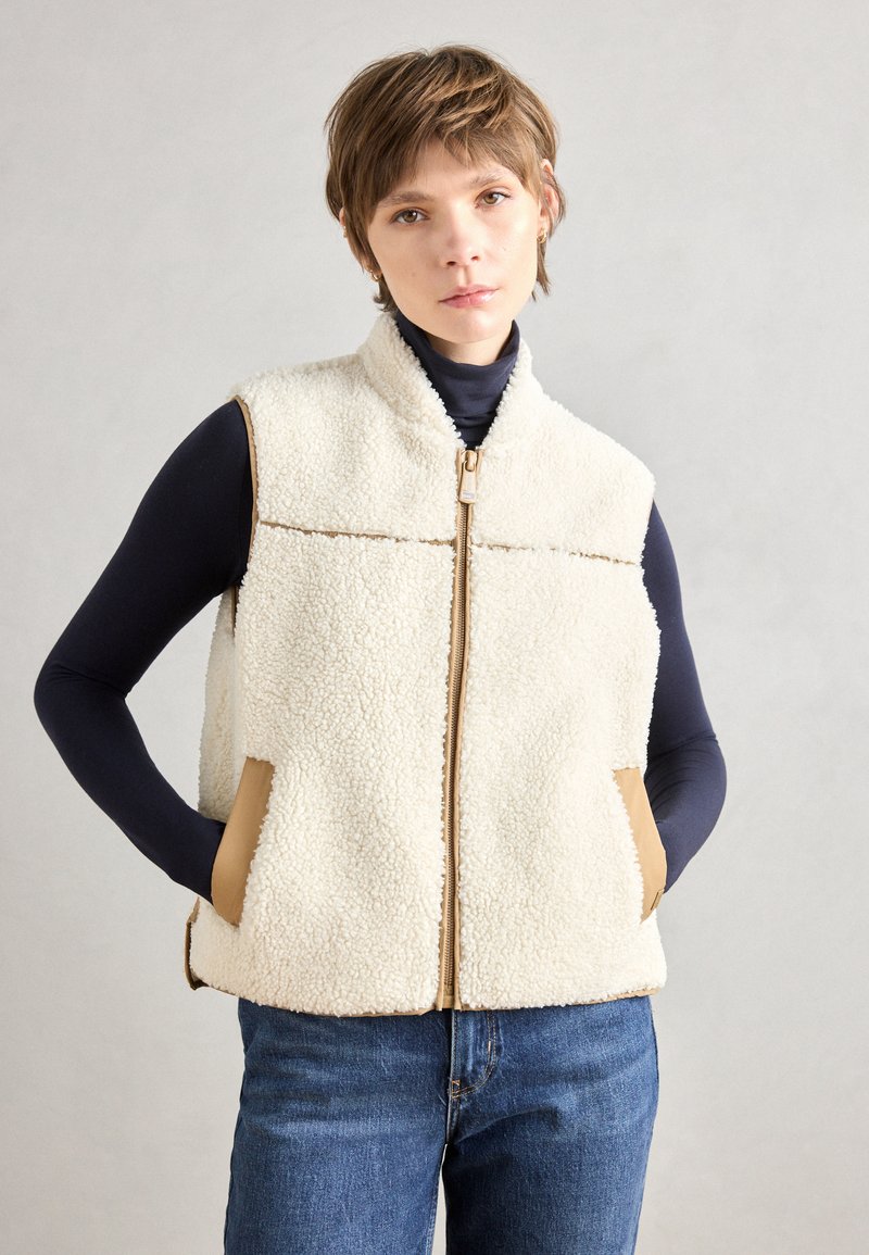Cream fleece vest with a high collar, front zipper, brown accent pockets, and casual fit. Paired with a dark turtleneck and blue jeans.