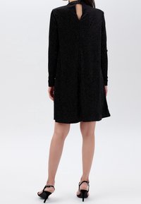Černé šaty s dlouhým rukávem a bílými puntíky, s detailem ve tvaru klíčové díry na zádech, volným střihem a jemnou texturou. Nosené s černými vysokými podpatky.