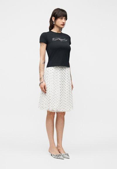 Γυναίκα όρθια, φοράει μαύρο τοπ με κοντά μανίκια με κείμενο «Perfect», λευκή φούστα μέχρι τα γόνατα με μικρό μαύρο σχέδιο και ασημένια μυτερά παπούτσια.