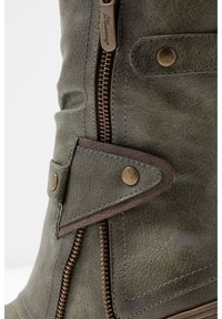 Botín verde con un zipper decorativo, botones metálicos y detalles de costura, que enfatizan su material texturizado y elementos de diseño.