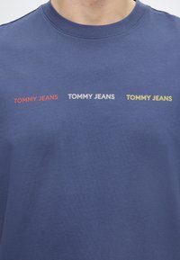 Μπλε T-shirt με στρογγυλή λαιμόκοψη με την επιγραφή «TOMMY JEANS» τυπωμένη τρεις φορές στο στήθος με κόκκινα, λευκά και κίτρινα γράμματα.