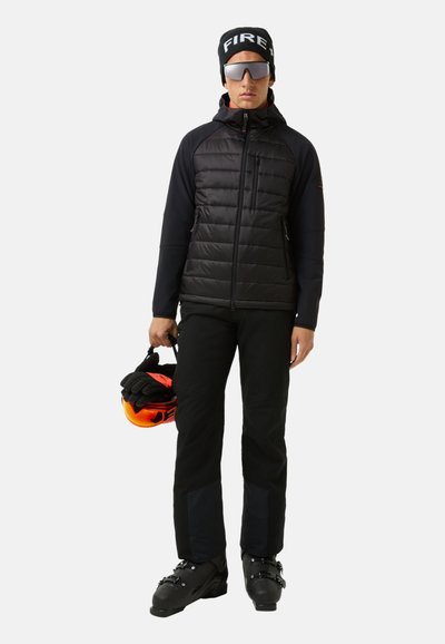 Sort polstret jakke med lynlås foran, kombineret med sorte skibukser. Modellen holder en orange hjelm og bærer en sort hue og solbriller.