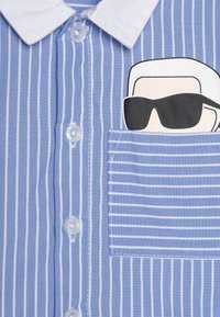 Chemise rayée bleu clair avec un col blanc, affichant le visage d'un personnage de dessin animé avec des lunettes de soleil sur la poche gauche. Six boutons.