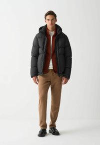 Jachetă puffer neagră cu glugă, strat interior ribbed portocalie, cămașă albă, pantaloni bej și pantofi negri. Texturi netede și culori contrastante.