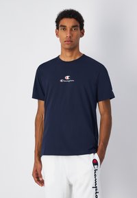 Camiseta de algodón azul marino con cuello redondo y mangas cortas. Presenta un logo "Champion" en rojo y blanco en el pecho. Diseño sencillo.