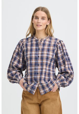 Femme aux cheveux blonds raides portant un chemisier à carreaux bleu et marron boutonné avec des manches bouffantes et un pantalon marron, les mains dans les poches.