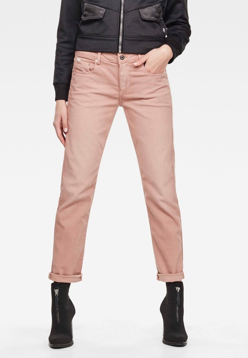 Žena oblečená v světle růžových zkrácených kalhotách, černé zipové bundě a černých kotníkových botách s předními zipy, stojící s jednou rukou v kapse.