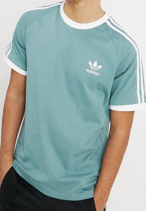 Mężczyzna w turkusowej koszulce Adidas z białym obszyciem i trzema białymi paskami na ramionach, ręce w kieszeniach czarnych spodni.