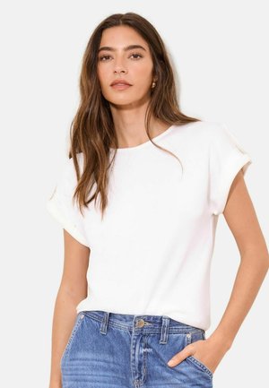 Mladá žena s dlhými hnedými vlasmi, oblečená v bielej krátkosleevej blúzke a modrých džínsoch, s jednou rukou v vrecku.