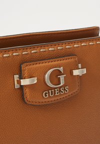 Brown textured leather handbag featuring a silver "G" logo and metal accent, with contrasting stitching on the edges of the design.