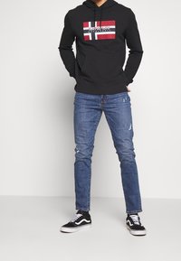 Czarna bluza z kapturem z czerwono-białą grafiką flagi Norwegii na przodzie, zestawiona z niebieskimi, przetartymi dżinsami oraz czarnymi trampkami Vans.