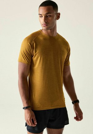 Mann in senfgelbem kurzärmligen Hemd und schwarzen Shorts steht mit dem Kopf zur Seite gedreht vor einfachem hellem Hintergrund.