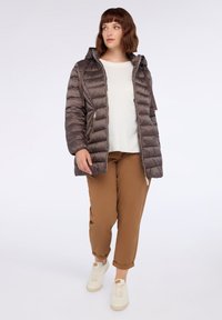 Chaqueta acolchada de color marrón oscuro con capucha, diseño acolchado, bolsillos con cremallera y silueta ajustada. Combinada con una blusa color crema y pantalones marrones.