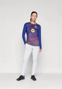 Camisola desportiva azul de manga comprida com riscas horizontais multicoloridas e logótipo, combinada com calças brancas e sapatilhas desportivas pretas.