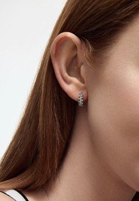 Srebrne kolczyki koła z teksturowaną powierzchnią i małymi lśniącymi akcentami, noszone na uchu modelki, podkreślające ich okrągły kształt.