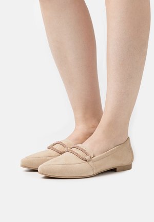 Mocassins en suède beige avec un détail tressé scintillant portés pieds nus, présentés sur un fond blanc uni.