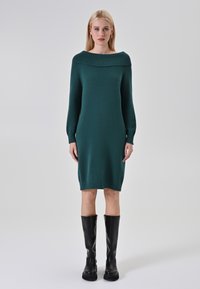 Abito in maglia teal con scollatura off-the-shoulder, maniche lunghe e polsini a coste; abbinato a stivali alti in pelle nera.