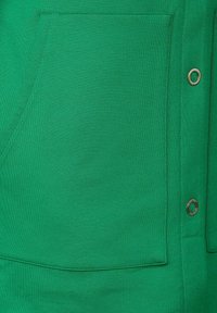 Indumento in tessuto verde con tasca applicata e rivetto metallico rotondo. Tessuto liscio con dettagli di cucitura sottili.