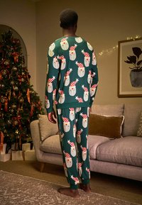 Žalias kalėdinis pižamos komplektas su pasikartojančiu Santa Claus veidų raštu, kuris puošia raudonas ir baltas skrybėles. Minkšta medžiaga; ilgomis rankovėmis ir kelnėmis.