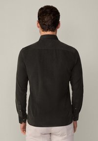 Camisa verde oscuro de manga larga, con detalle de botones en los puños, cuello clásico y una tela texturizada, vista desde la parte de atrás.