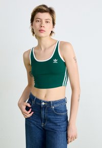 Grön crop top med vit kant och tre vita ränder på sidorna, med en Adidas-logotyp. Matchad med högmidjade blå jeans.