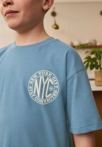 Lyseblå bomulds-T-shirt med en cirkulær grafik med teksten "New York City" og "Stay Connected" i cremefarve. Kortærmet, rund halsudskæring.