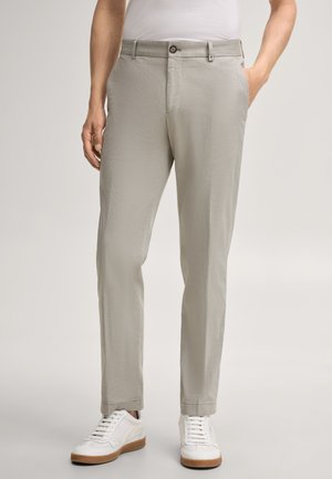 Mężczyzna ubrany w jasnoszare obcisłe spodnie i białe trampki, stoi z rękami w kieszeniach na jednolitym tle.