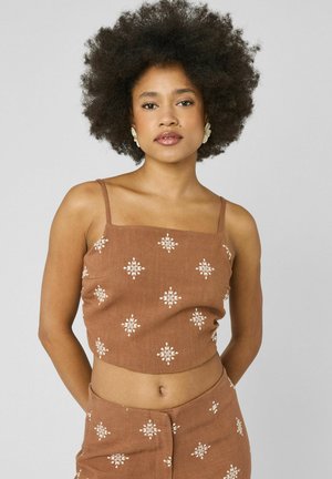 Femme aux cheveux courts et bouclés portant un crop top marron et un pantalon assorti avec des motifs floraux blancs brodés, debout devant un fond uni.