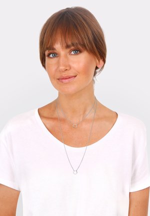 Silver necklaces with circular pendants, layered over a white t-shirt. The shorter chain features a small ring, while the longer chain holds a larger ring.