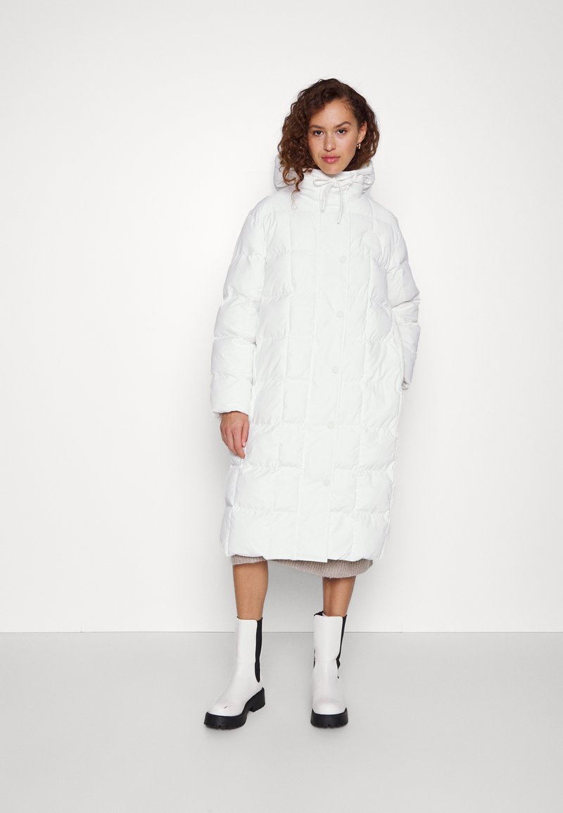 Manteau matelassé blanc avec capuche, présentant un design matelassé, de gros boutons et une coupe jusqu'aux genoux. Porté par-dessus un vêtement en tricot beige et des bottes blanches.