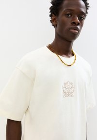 Cream-colored cotton T-shirt, short sleeves, featuring a floral embroidered design in brown on the chest, round neckline.